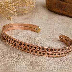 May include: A copper cuff bracelet with a hammered texture and two rows of evenly spaced circular cutouts. The bracelet has a warm, reddish-brown hue and a simple, open design. The bracelet is approximately 2.5 inches wide.