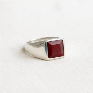 May include: A silver ring with a large, square, deep red gemstone. The ring has a simple, modern design with a polished finish. The gemstone is set in a square bezel, and the band is smooth and wide.