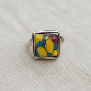 May include: A square silver ring featuring a vibrant stone with yellow, blue, and red hues. The stone is set within a silver bezel, creating a striking contrast. The ring's design is simple yet eye-catching, suitable for everyday wear.
