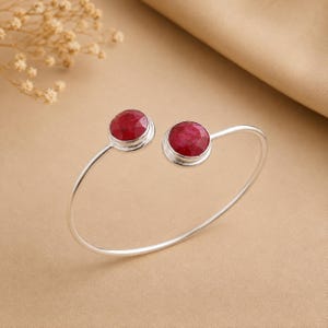 Può includere: Un bracciale a fascia aperta in argento con due pietre preziose di rubino rosso scuro, rotonde e sfaccettate, incastonate a ciascuna estremità. Il cinturino in metallo è liscio e lucido, creando una forma circolare aperta sul davanti.