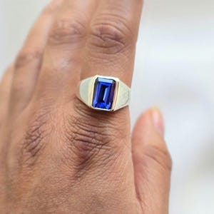 Könnte beinhalten: Ein silberner Siegelring mit einem rechteckigen, tiefblauen Edelstein. Der Ring hat einen klassischen Siegelstil mit einer goldfarbenen Fassung um den Edelstein. Der Ring wird an einem Finger gezeigt.