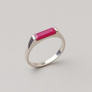Peut inclure: Une bague en argent ornée d'une pierre rouge vif de forme rectangulaire. La bague présente un design simple et moderne avec une finition polie. La pierre est sertie horizontalement dans la monture.