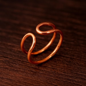 Könnte beinhalten: Ein kupferfarbener Ring mit einem einzigartigen, offenen Design. Der Ring hat ein Doppelband mit einer schleifenförmigen, abstrakten Form oben. Er liegt auf einer dunklen Holzoberfläche, die seinen metallischen Glanz hervorhebt.