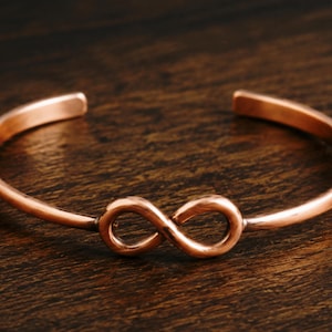 Op de afbeelding: Een koperkleurige manchetarmband met een oneindigheidssymbool in het midden. De open armband is gemaakt van één stuk metaal en wordt gepresenteerd op een donkere houten ondergrond.