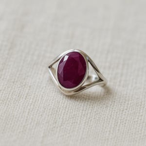 May include: A silver ring featuring an oval, deep red gemstone. The ring's band has a unique open design on either side of the gemstone, creating a modern and elegant look. The gemstone is the focal point, showcasing its rich color and cut.