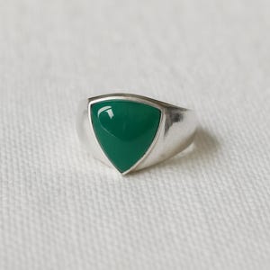 May include: A silver ring with a triangular, emerald-green stone. The ring has a smooth, polished finish and a wide band. The gemstone is set flush with the ring's surface, creating a sleek, modern look. The ring is displayed on a white, textured surface.