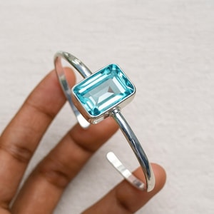 May include: A silver cuff bracelet with a rectangular, light blue gemstone. The gemstone is set in a square bezel. The open-ended bracelet is a simple, elegant design. The gemstone is a focal point.