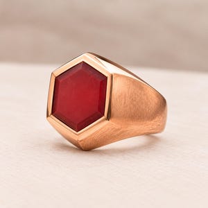 May include: A rose gold ring featuring a prominent hexagonal-shaped red stone set into the center of the band. The metal surface of the ring has a brushed texture, and the red stone is flat with beveled edges, creating a geometric design.