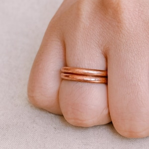 May include: Two copper-colored rings are stacked on a finger. The rings have a smooth, rounded design and a warm, metallic hue. The background is a soft, neutral color, highlighting the rings' simple elegance. The rings are a classic jewelry item.
