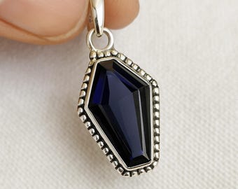 May include: A silver pendant with a dark blue, coffin-shaped gemstone. The gemstone is faceted and set within a beaded border. The pendant has a small loop at the top for a chain.