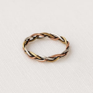 May include: A braided ring with interwoven strands of silver, gold, and copper-toned metal. The ring has a textured, woven appearance against a soft, white background. This piece of jewellery is a unique accessory.