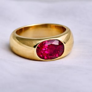 May include: A gold ring with a large, oval-cut, ruby-red gemstone. The ring has a thick, rounded band and a polished finish. The gemstone is set in a bezel setting, and the ring is displayed on a white surface.