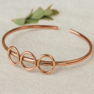 Peut inclure: Un bracelet manchette ouvert de couleur cuivrée. Le bracelet comporte trois cercles reliés au centre. Le bijou est présenté sur une surface de couleur neutre, avec une branche de feuilles vertes en arrière-plan.