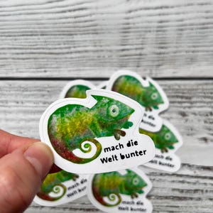 Könnte beinhalten: Mehrere grüne, gelbe und rote Chamäleon-Aufkleber. Jeder Aufkleber zeigt ein Chamäleon mit dem Text "mach die Welt bunter" in einer weißen Sprechblase. Die Aufkleber befinden sich auf einer hellen Holzmaserung.
