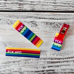 Könnte beinhalten: Drei rechteckige Stoffetiketten mit einem leuchtenden Regenbogen-Streifenmuster. Jedes Etikett zeigt den weißen Text FCK AFD, der deutlich auf der Oberfläche aufgedruckt ist. Ein Etikett ist an einer roten Kunststoff-Nähklammer befestigt, zwei liegen flach.