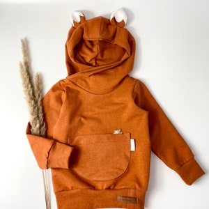 May include: A brown hooded sweatshirt with fox ears, a kangaroo pocket, and a small bear applique. The sweatshirt has a brown label with the text "Its Lovely" on the bottom.