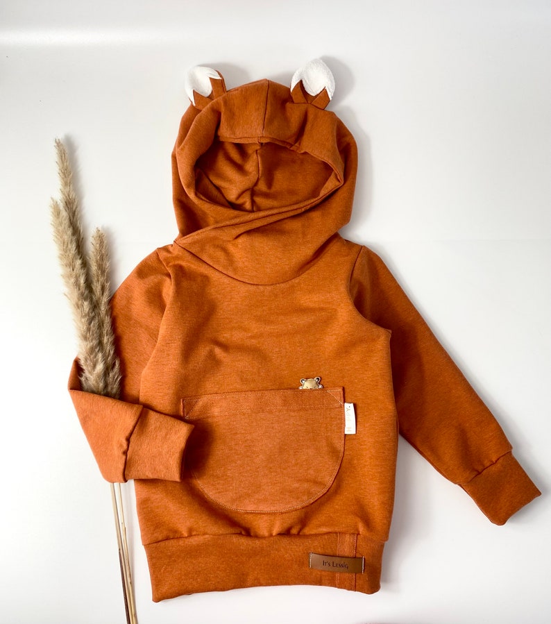 May include: A brown hooded sweatshirt with fox ears, a kangaroo pocket, and a small bear applique. The sweatshirt has a brown label with the text "Its Lovely" on the bottom.