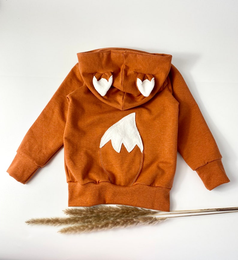 May include: A brown hooded sweatshirt with a white fox tail and ears on the back. The sweatshirt is made of a soft, comfortable fabric and has a relaxed fit.