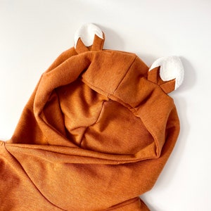 May include: A brown hooded sweatshirt with white fox ears. The hood is pulled up and the sweatshirt is folded in half.