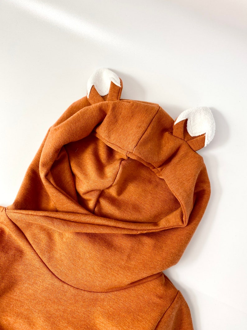 May include: A brown hooded sweatshirt with white fox ears. The hood is pulled up and the sweatshirt is folded in half.