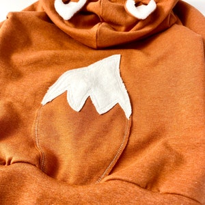 May include: A brown hooded sweatshirt with a white fox tail and ears. The sweatshirt is made of a soft, comfortable fabric.