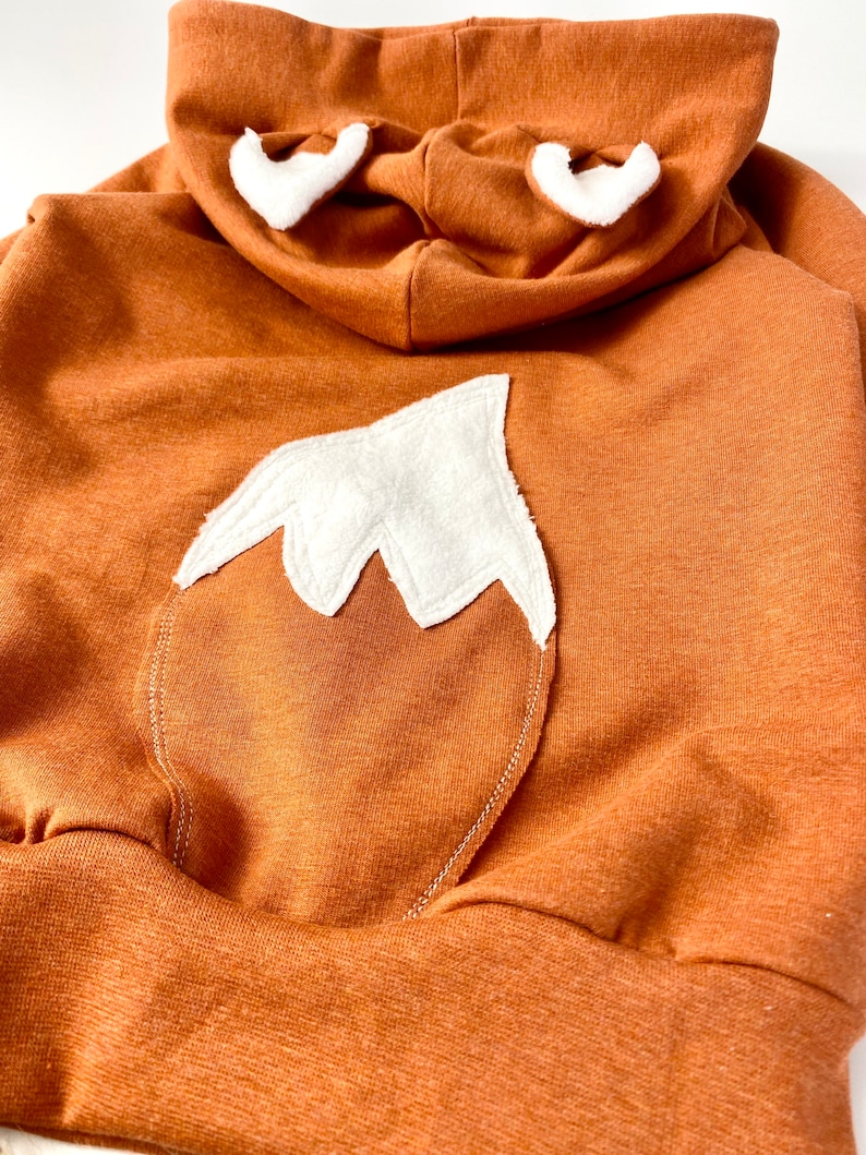 May include: A brown hooded sweatshirt with a white fox tail and ears. The sweatshirt is made of a soft, comfortable fabric.