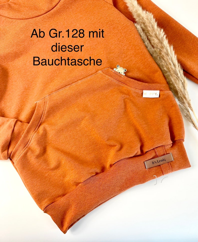 May include: A rust-colored hoodie with a kangaroo pocket and a small brown bear applique. The hoodie has a ribbed hem and cuffs. The text "Ab Gr. 128 mit dieser Bauchtasche" is printed on the front of the hoodie. The text "It's Lessic" is printed on a brown label sewn to the bottom of the hoodie.