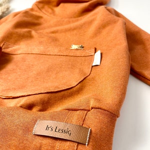 May include: A rust-colored hooded sweatshirt with a kangaroo pocket and a brown leather patch that says "It's Lessig".