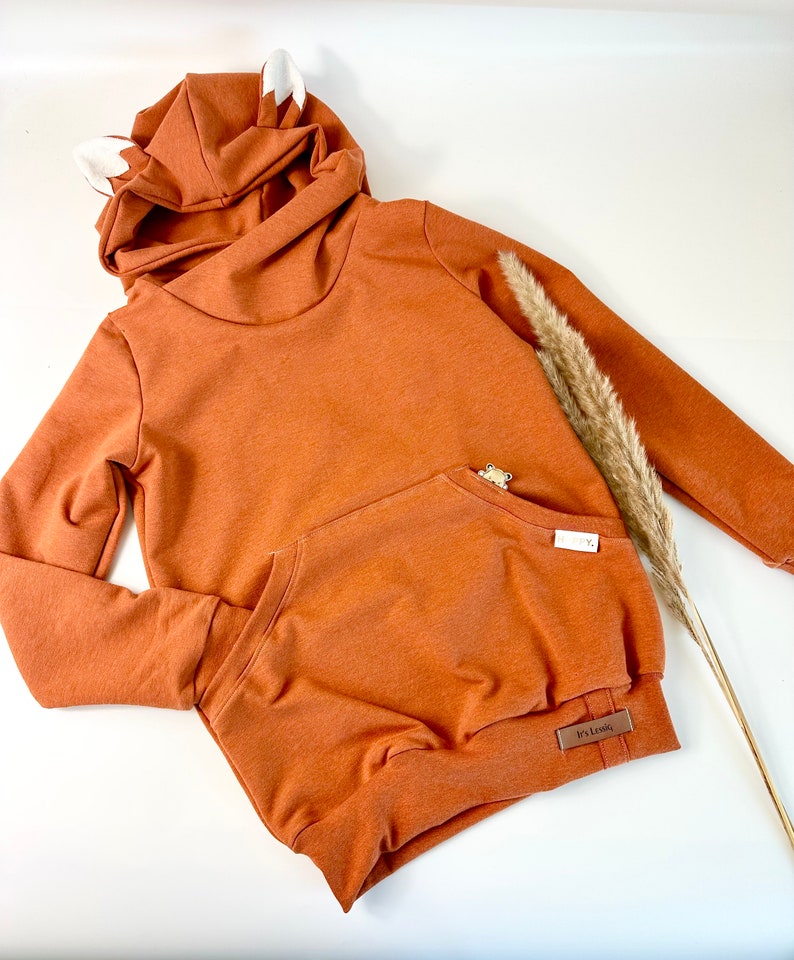 May include: A rust-colored hooded sweatshirt with a kangaroo pocket and fox ears on the hood. The sweatshirt has a brown tag that says "It's Lovely".