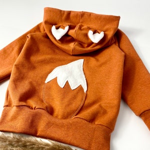 May include: A brown hooded sweatshirt with a white fox tail and ears on the back. The sweatshirt has a ribbed hem and cuffs.