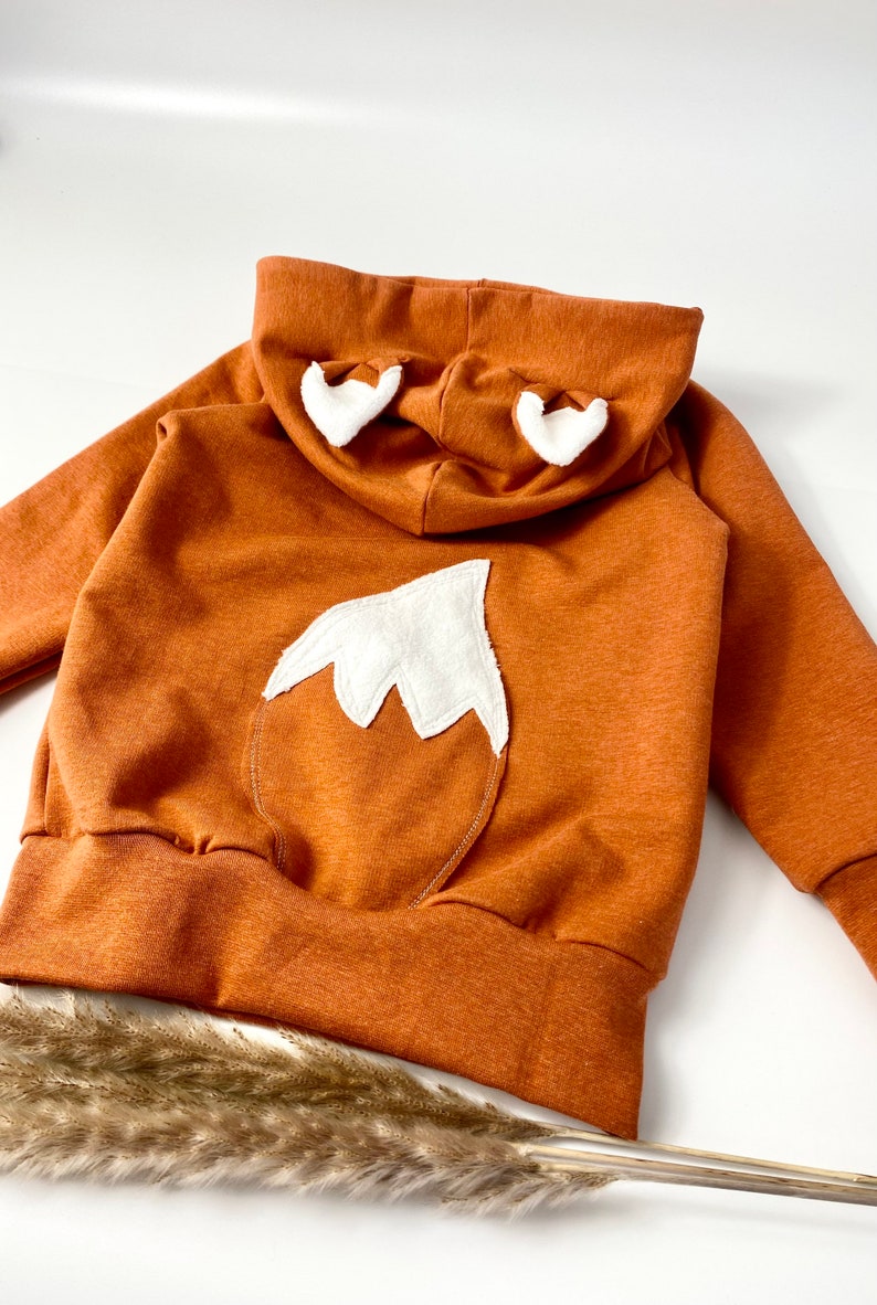May include: A brown hooded sweatshirt with a white fox tail and ears on the back. The sweatshirt has a ribbed hem and cuffs.