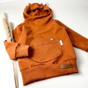 May include: A brown hooded sweatshirt with a kangaroo pocket and a small bear applique. The sweatshirt has a fold-over collar and a label that says "It's Lesslie".