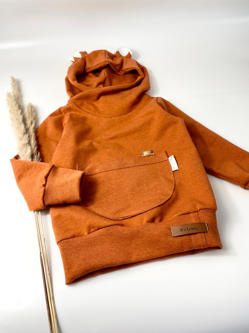 May include: A brown hooded sweatshirt with a kangaroo pocket and a small bear applique. The sweatshirt has a fold-over collar and a label that says "It's Lesslie".