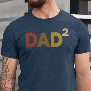 May include: A navy blue t-shirt with the text "DAD²" in a retro-style font with red, yellow, and orange colors.