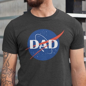 May include: A dark heather grey t-shirt with a NASA logo featuring the word "DAD" in white text.