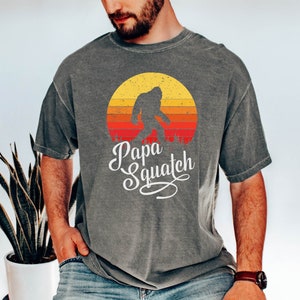 May include: A gray t-shirt with a vintage-style graphic of a Bigfoot walking in front of a sunset. The text "Papa Squatch" is written below the graphic.