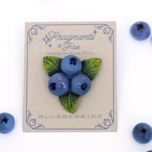 Puede incluir: Un pin hecho a mano con tres arándanos azules y hojas verdes. El pin está en una tarjeta con el texto "Fragments of Fae Handmade Pin Blueberries".
