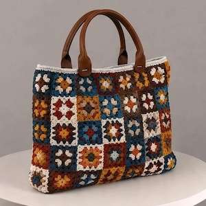 Può includere: Una borsa tote realizzata con toppe all'uncinetto granny square multicolori nei toni del marrone, blu, bianco e giallo. La borsa presenta due manici in similpelle marrone fissati sul bordo superiore, conferendo una forma rettangolare.