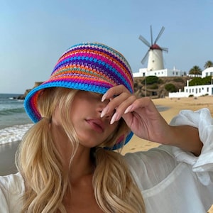 May include: A colorful crochet bucket hat with a blue brim. The hat features horizontal stripes in shades of blue, pink, orange, and purple. The hat is worn on a sunny day at the beach. The background includes a windmill and the ocean.