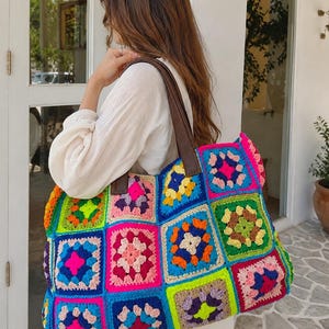 Peut inclure: Un grand sac fourre-tout composé de carrés de crochet multicolores dans des tons vifs de rose néon, bleu électrique, vert citron, orange et jaune. Le sac est doté de deux bandoulières en cuir marron foncé fixées sur le bord supérieur.