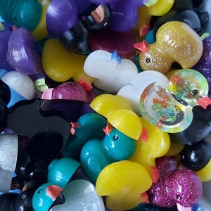 May include: A collection of colourful rubber duck figurines in various colours, including purple, teal, yellow, white, black, and glitter. The ducks are arranged in a pile, with some overlapping.