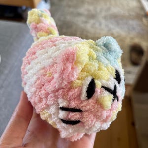Könnte beinhalten: Ein kleines, rundes Plüschkatzenspielzeug aus weichem, buntem Garn in Pastelltönen wie Rosa, Gelb, Weiß und Hellblau. Das Stofftier verfügt über gestickte schwarze Faden-Details für Augen, Nase und Schnurrhaare sowie einen kleinen Schwanz.