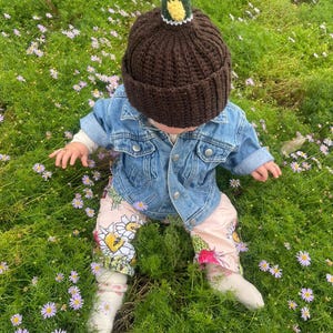 May include: A dark brown ribbed knit beanie featuring a plush mallard duck head on top, worn with a light blue denim jacket and light pink pants decorated with a colorful floral and cactus print, paired with white socks.