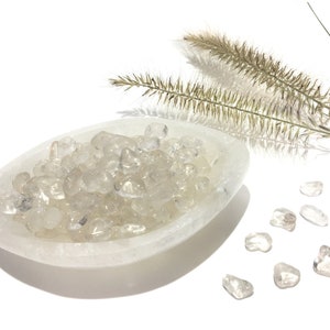 Könnte beinhalten: Eine weiße Selenit-Schale gefüllt mit klaren Quarzkristallen. Die Schale steht auf einer weißen Oberfläche. Es liegen auch einige lose klare Quarzkristalle um die Schale herum.