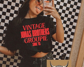 May include: A black short-sleeved t-shirt featuring red text that reads VINTAGE JONAS BROTHERS GROUPIE SINCE '05. The shirt is paired with black distressed denim shorts. A black baseball cap and brown-tinted sunglasses are also worn with the outfit.