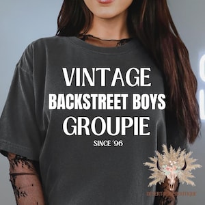 Puede incluir: Camiseta gris oscuro con las palabras "VINTAGE BACKSTREET BOYS GROUPIE" en blanco. Debajo, "SINCE '96". La camiseta es de estilo vintage, probablemente de algodón suave.