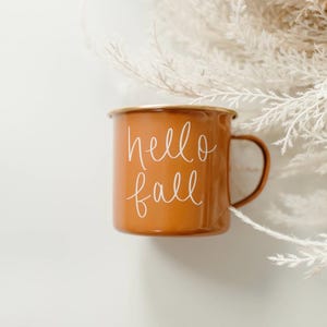 May include: An orange enamel mug with the words "hello fall" in white script. The mug has a brown handle and rim. White feathery decorative elements are in the background.