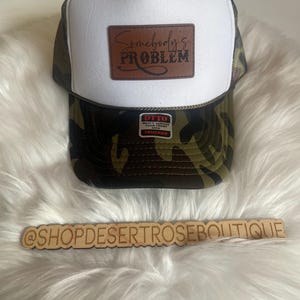 Puede incluir: Gorra de camionero blanca y de camuflaje con un parche de cuero marrón que dice "Somebody's Problem". La gorra también tiene una etiqueta roja que dice "Otto World Famous Foam Front Trucker".