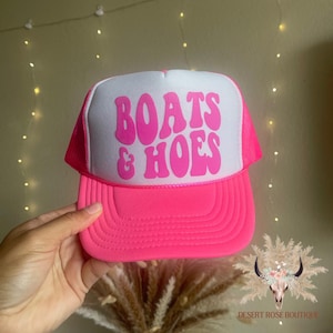 Könnte beinhalten: Rosa-weiße Trucker-Kappe mit dem Text "BOATS & HOES" in rosa Buchstaben.