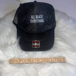 May include: Black trucker hat with a mesh back and embroidered black butterflies and a heart with the text "ALL BLACK EVERYTHING" in white. The hat has a black Otto trucker label.